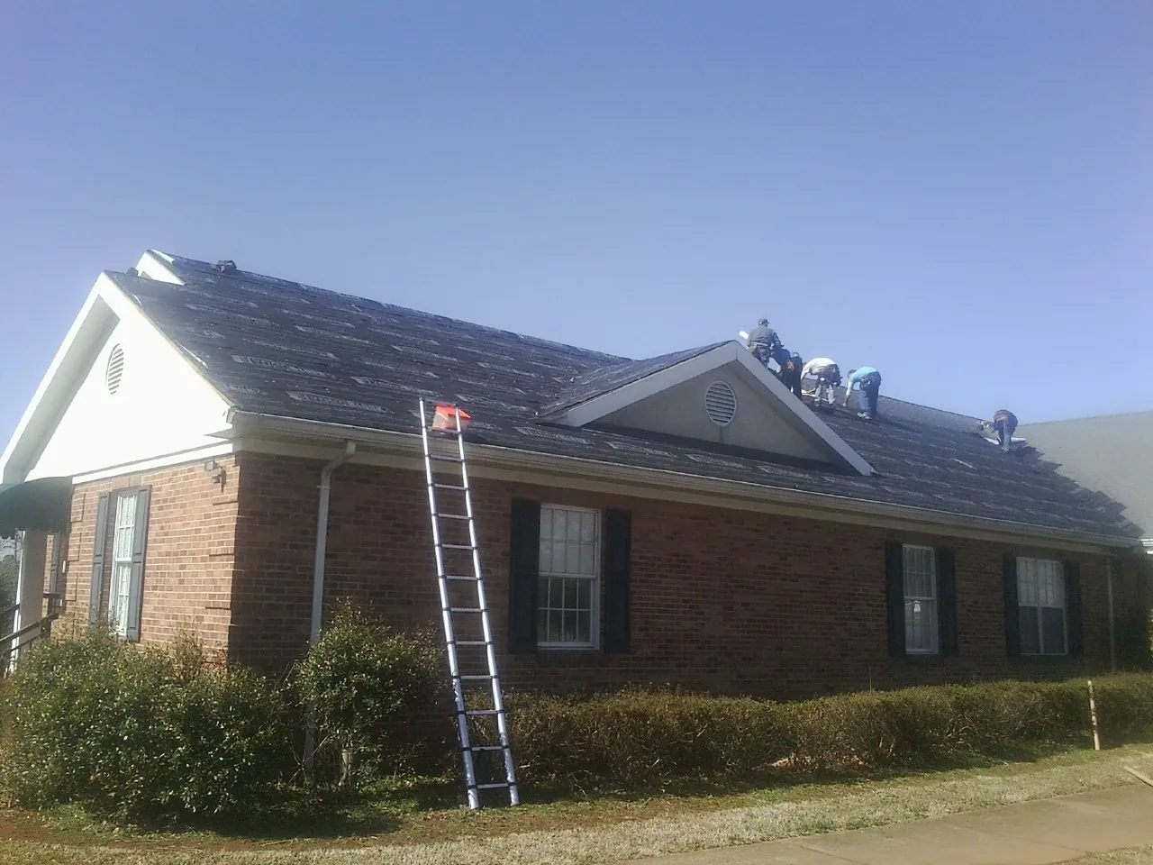 Crew tearing shingles off a brick ranch roof with a ladder set — Emergency Roof Repair work by our crew in Sunset