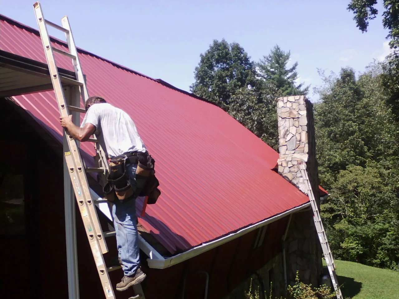 Roofer on a ladder beside a red metal roof and stone chimney — Soffit & Fascia Repair by Roofing Brothers in Sunset